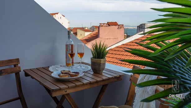 Ein Holztisch mit einer Auswahl an Gebäck, einer Weinflasche und einem Glas auf einem Balkon mit Blick auf eine Stadtlandschaft mit Terrakotta-Dächern und blauem Himmel.