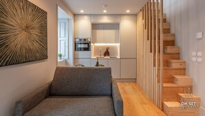 Ein modernes Wohnzimmer mit grauem Sofa, einer Holztreppe und einer Küchenzeile inklusive Einbauofen sowie einem Weinglas auf der Arbeitsfläche.
