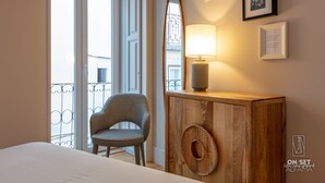 Ein Schlafzimmer mit einem Bett, einem Sessel, einer Holzkommode mit Spiegel und Lampe sowie einem Balkon mit Geländer.