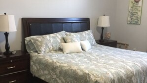 A neatly made bed with a patterned bedspread and pillows, flanked by two bedside tables with lamps, in a room with a plain wall and a decorative item hanging above the bed. - Ocean Front, Luxurious 3/3 New Rental (Daytona Beach Shores)