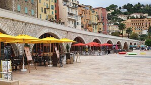 An outdoor café with stone arches, yellow and red umbrellas, and a stone building in the background. - Beautiful 3 pieces in luxury residence 50 meters from the sea (Menton)