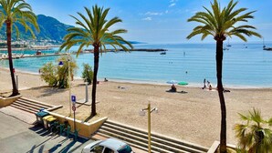 A beach with palm trees, a clear sky, and boats in the water. - Beautiful 3 pieces in luxury residence 50 meters from the sea (Menton)