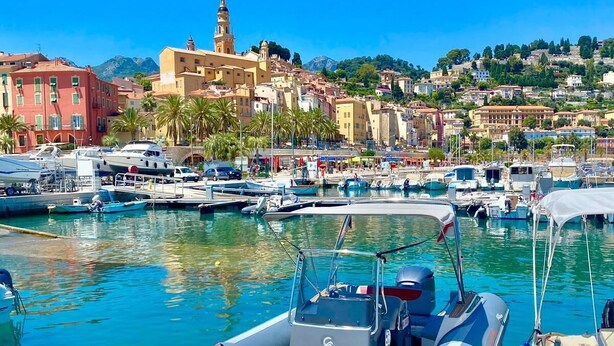 A marina with boats docked, a prominent building with a tower, and a hillside with buildings and greenery in the background. - Beautiful 3 pieces in luxury residence 50 meters from the sea (Menton)