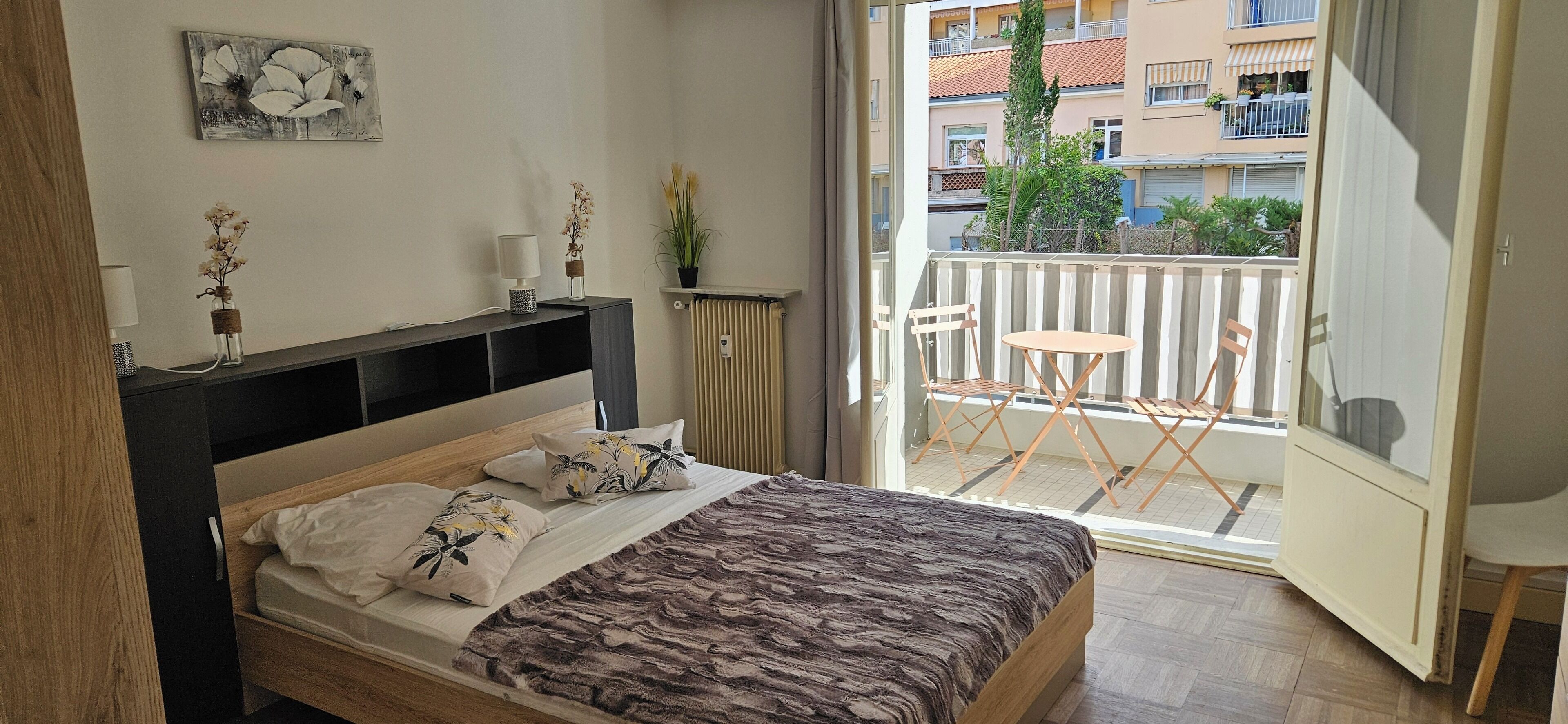 A bedroom with a bed, bedside table, lamp, and a view of a balcony with chairs and a table.