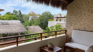 A balcony with a wooden chair and a small table, a potted plant, and a view of buildings with thatched roofs and lush greenery.