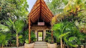 A thatched-roof structure with a sign reading "Natura" in a lush, tropical garden setting.