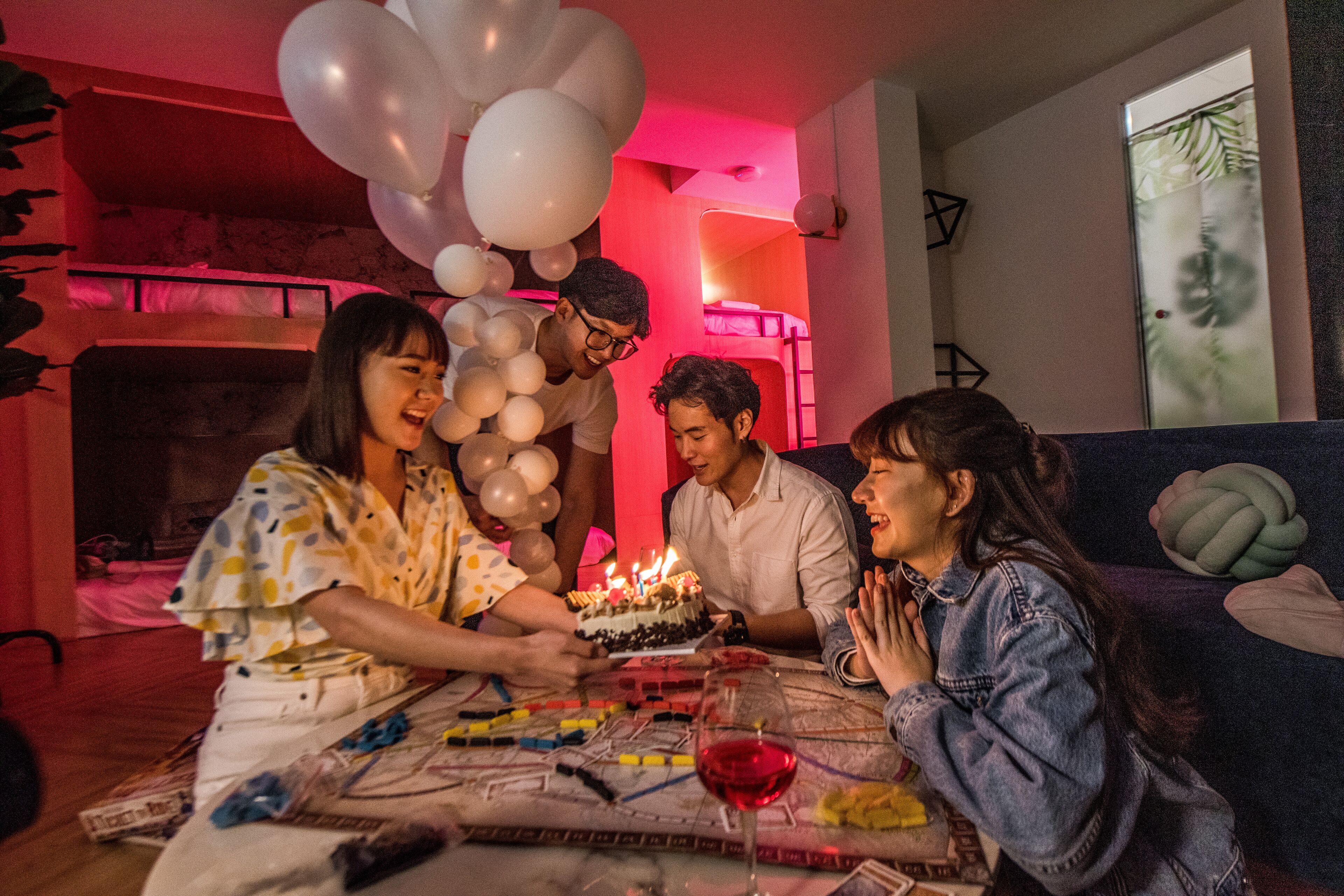 A group of people celebrating a birthday with a cake and balloons in a room with bunk beds and a window with a plant.