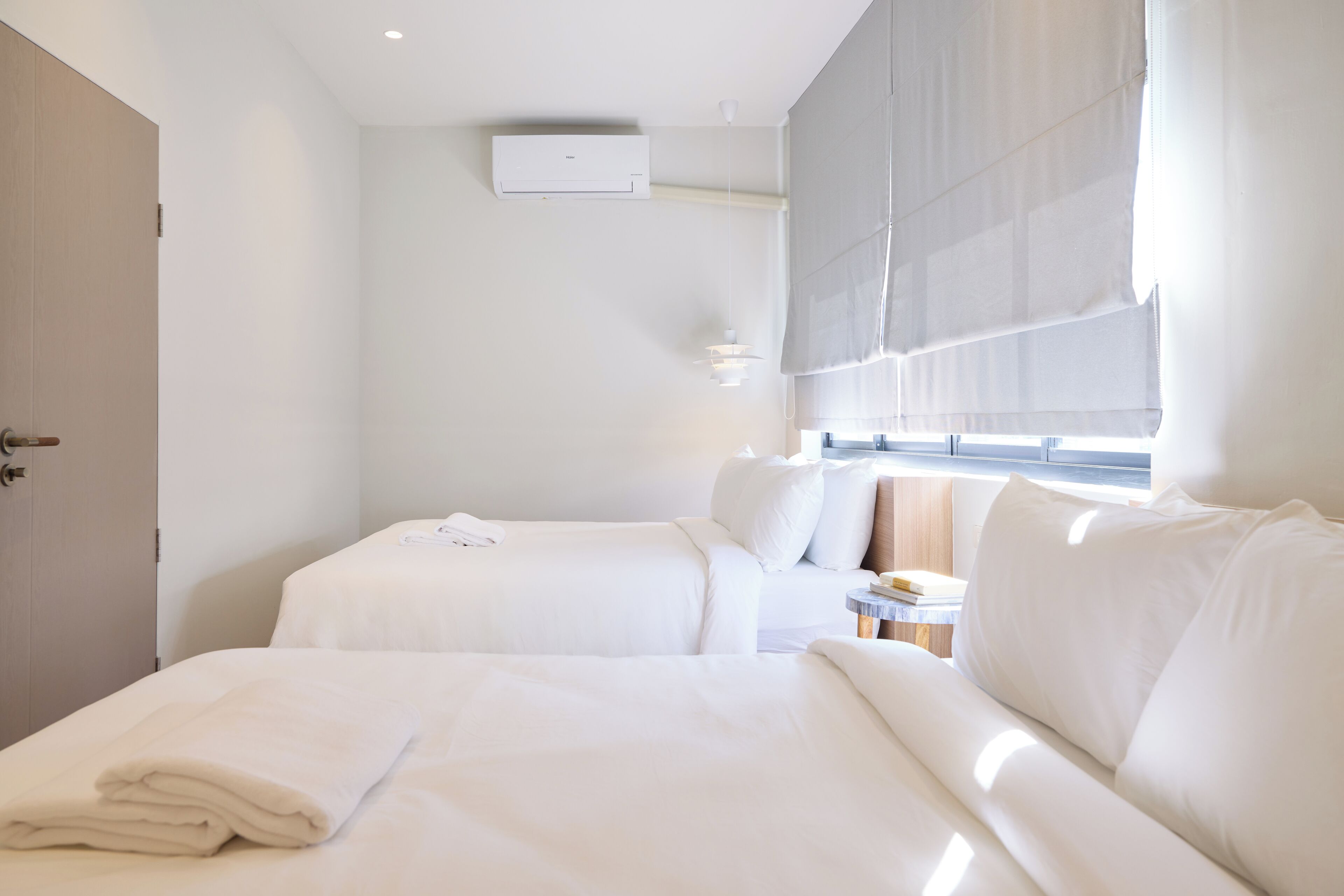 A hotel room with two beds, a bedside table, a window with blinds, an air conditioning unit, and a door.