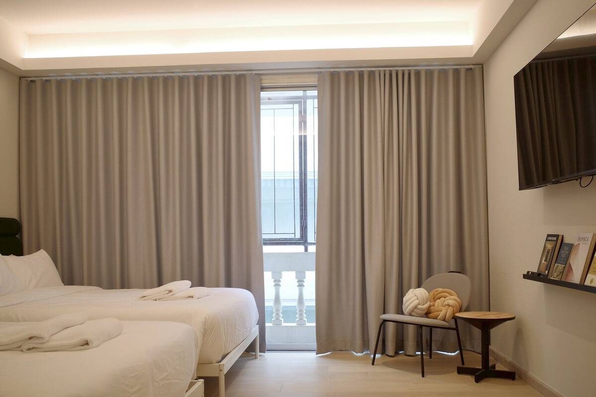 Habitació d'hotel amb dos llits, una cadira, una taula petita, un televisor i un balcó amb cortines.