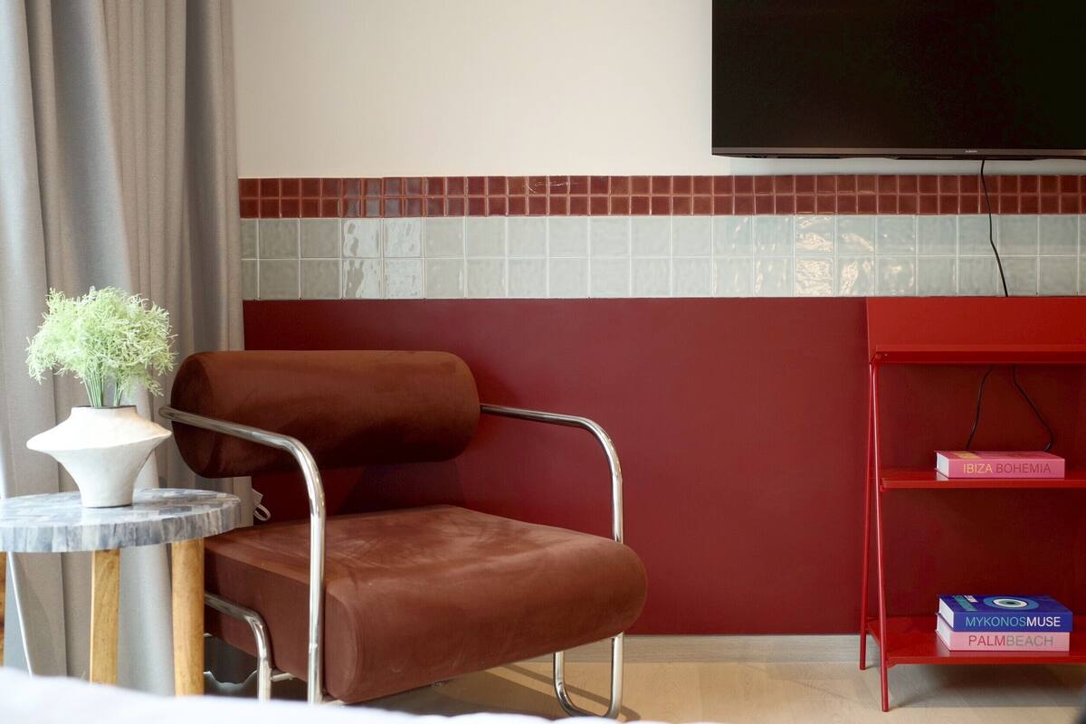 Una habitació d'hotel moderna amb una cadira de pell marró, una taula rodona petita amb un vas de flors blanc, una prestatgeria vermella amb llibres i un rebedor revestit de rajoles.