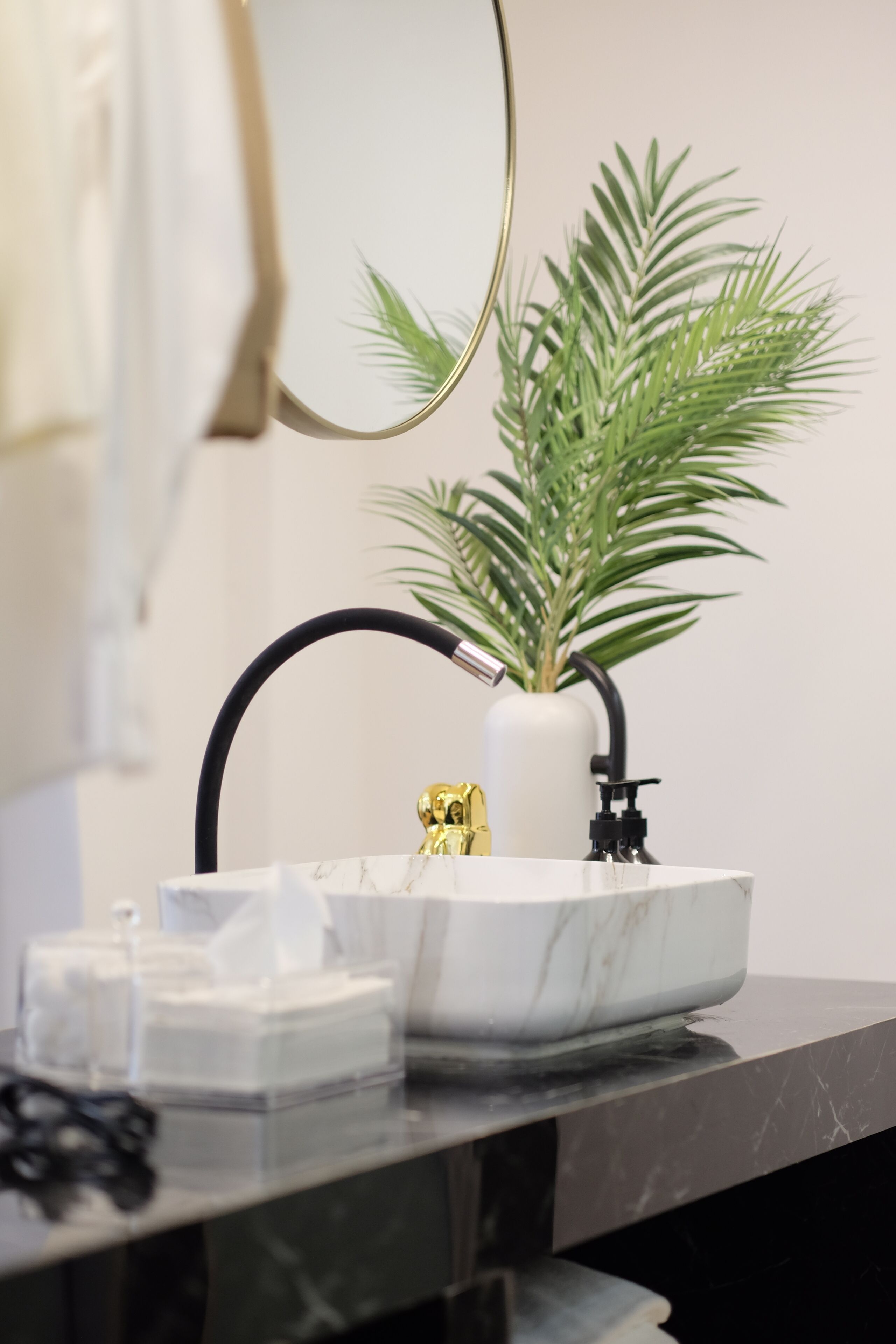 A bathroom with a marble sink, a gold faucet, a black hose, a white vase with a plant, and a mirror.