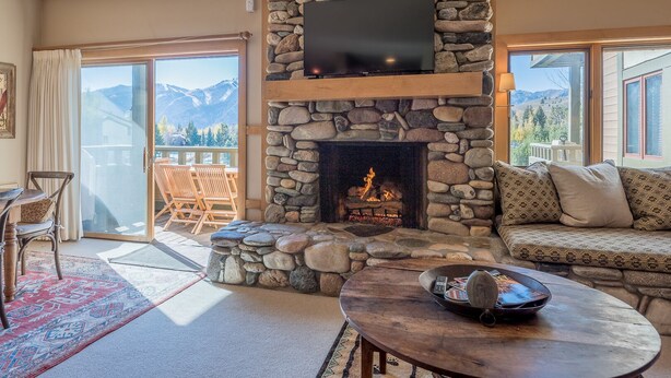 Una chimenea de piedra con un televisor de pantalla plana arriba, una mesa de madera con una bandeja, un sofá estampado y vistas a las montañas a través de amplios ventanales.