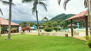 A resort with a swimming pool, palm trees, and a mountain backdrop. - House in Ubatuba - Gated community | swimming pool | gourmet area | river access (Horto (Morro das Moças))