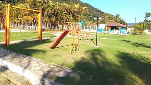 A playground with various play structures, a small building, and a grassy area with palm trees. - House in Ubatuba - Gated community | swimming pool | gourmet area | river access (Horto (Morro das Moças))