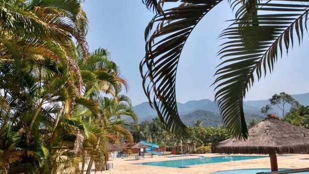 A pool area with palm trees and a thatched-roof structure. - House in Ubatuba - Gated community | swimming pool | gourmet area | river access (Horto (Morro das Moças))