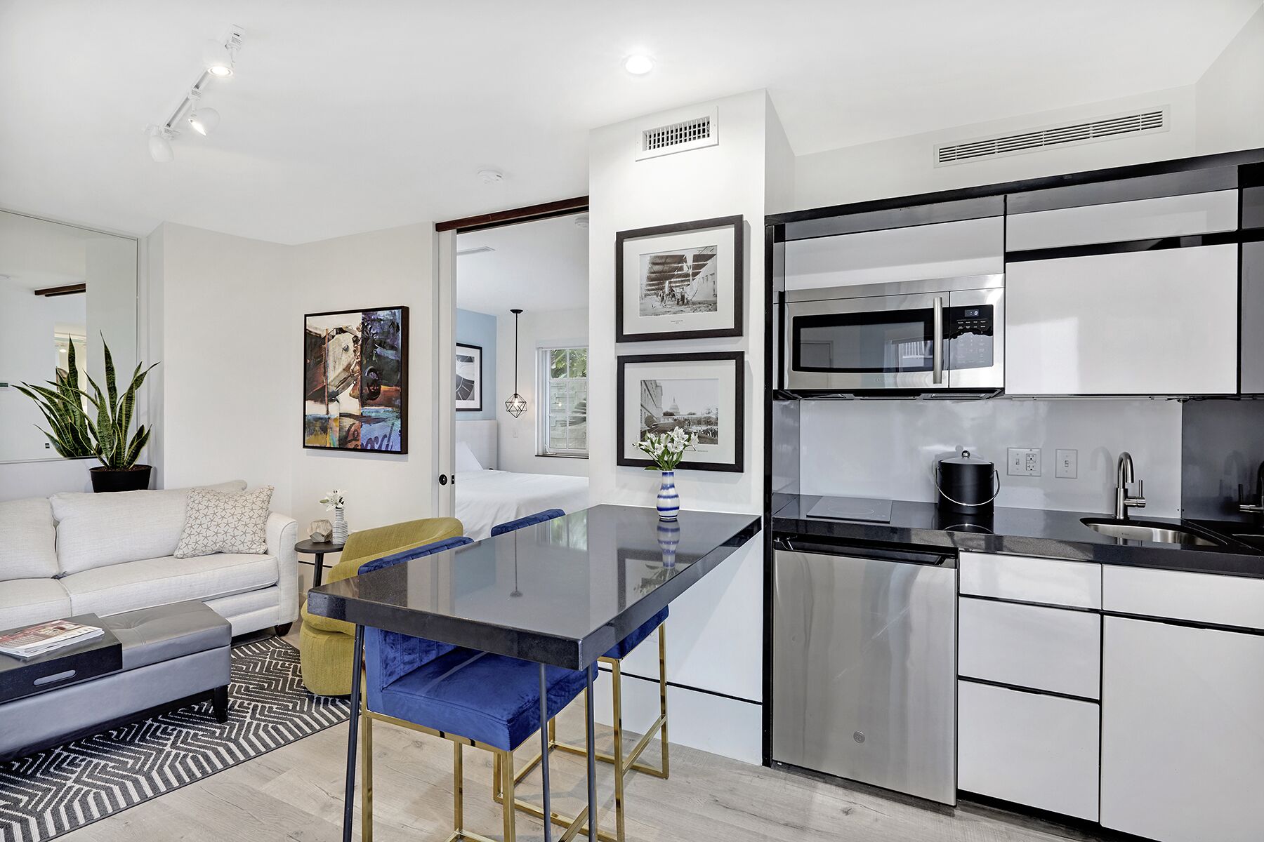 A modern kitchen with stainless steel appliances, a dining area with a glass table, and a living room with a sofa and a potted plant.