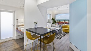 A modern kitchen with a black countertop and yellow chairs, a dining area with a sofa and a view of the sea, and a balcony with outdoor seating. - Ocean Treasure Beachside Suites (Lauderdale-by-the-Sea)