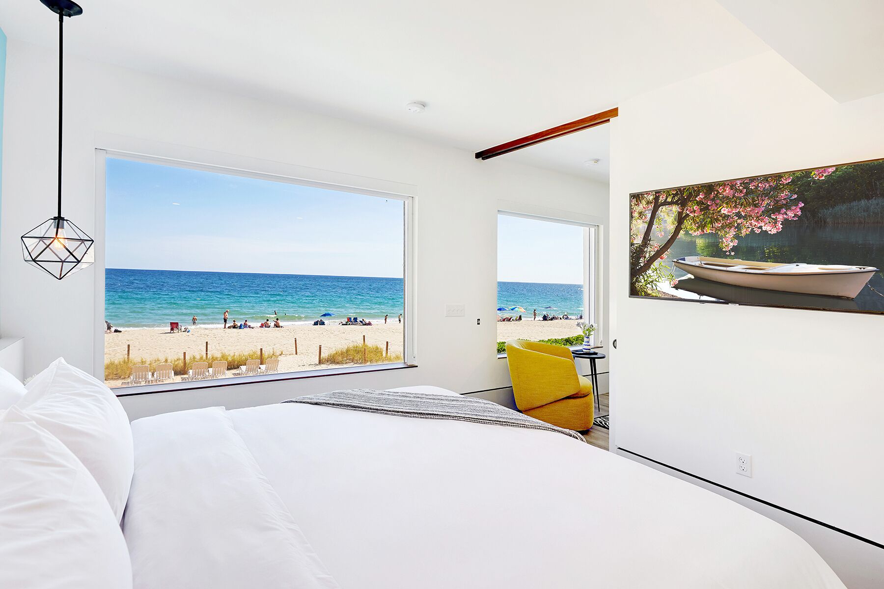 A bedroom with a large window overlooking a beach, a bed with white linens, a yellow chair, and a painting of a boat on the wall.