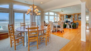 A spacious living area with a large dining table, comfortable seating, a kitchen area, and large windows offering a view of the ocean. - 2869 - Seascape - Oceanfront Duck Retreat w/ Pool, Hot Tub & Views (Kitty Hawk)