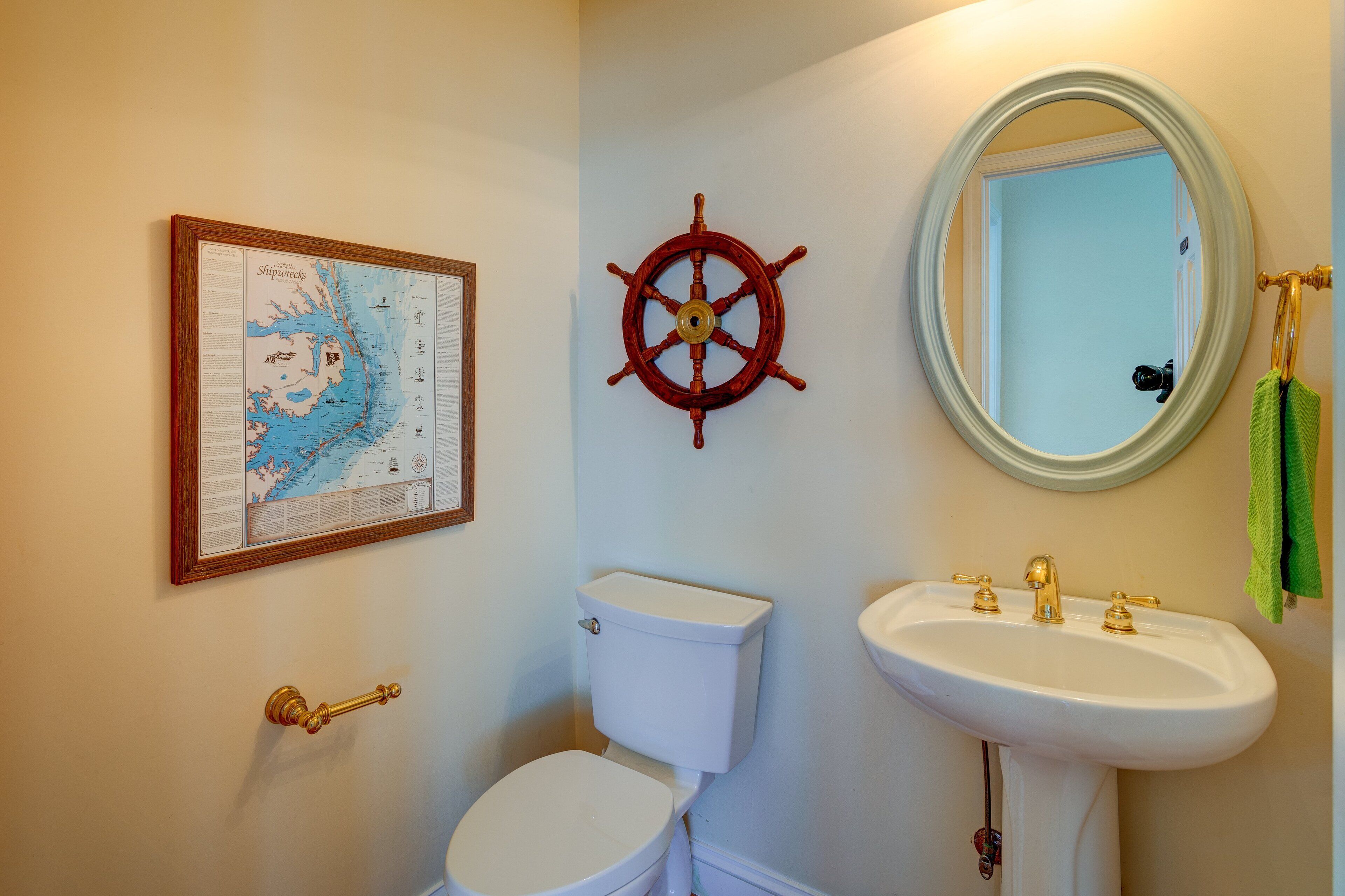 Un baño con un inodoro blanco, una rueda de barco, un mapa enmarcado y un lavabo con detalles dorados.