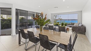 A modern dining area with a wooden table and black wicker chairs, a large window with a view of the sea, and a balcony with outdoor seating. - Maggie's Place Beachfront Retreat in Corlette (Corlette)