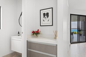 A modern bathroom with a white vanity, a framed black and white dog illustration, a vase with red flowers, and a view of the sea through a glass door. - Maggie's Place Beachfront Retreat in Corlette (Corlette)