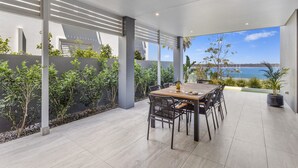 A modern outdoor dining area with a view of the sea, featuring a wooden table and metal chairs. - Maggie's Place Beachfront Retreat in Corlette (Corlette)