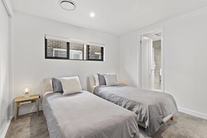 A bedroom with two beds, a window with blinds, a nightstand with a lamp, and a door leading to a bathroom. - Maggie's Place Beachfront Retreat in Corlette (Corlette)