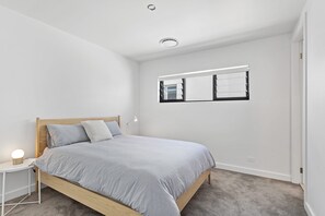A bedroom with a single bed, a bedside table with a lamp, a window with blinds, and a ceiling light.