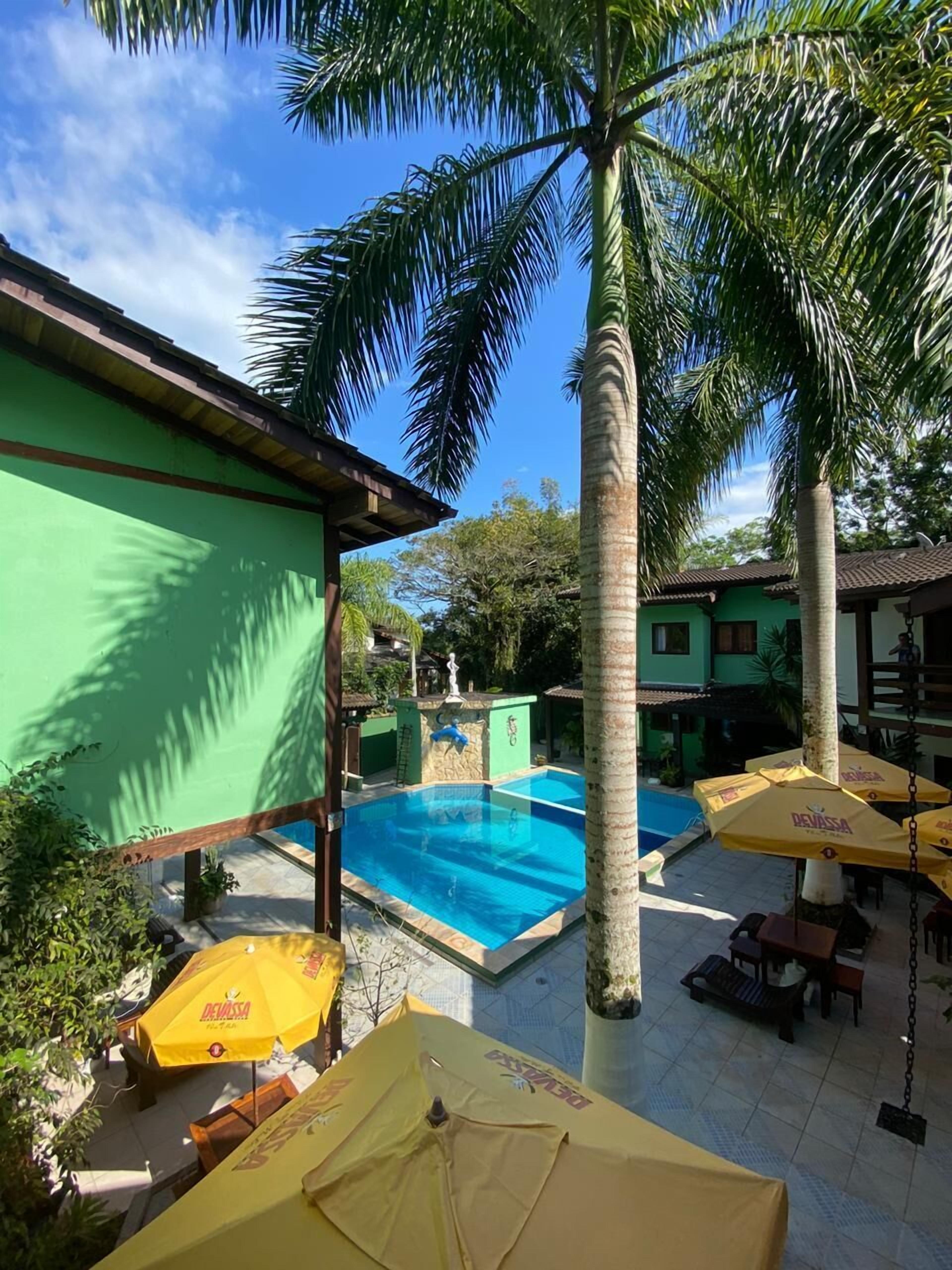 A pool area with a clear blue pool, surrounded by palm trees and green buildings. There are yellow umbrellas and lounge chairs.