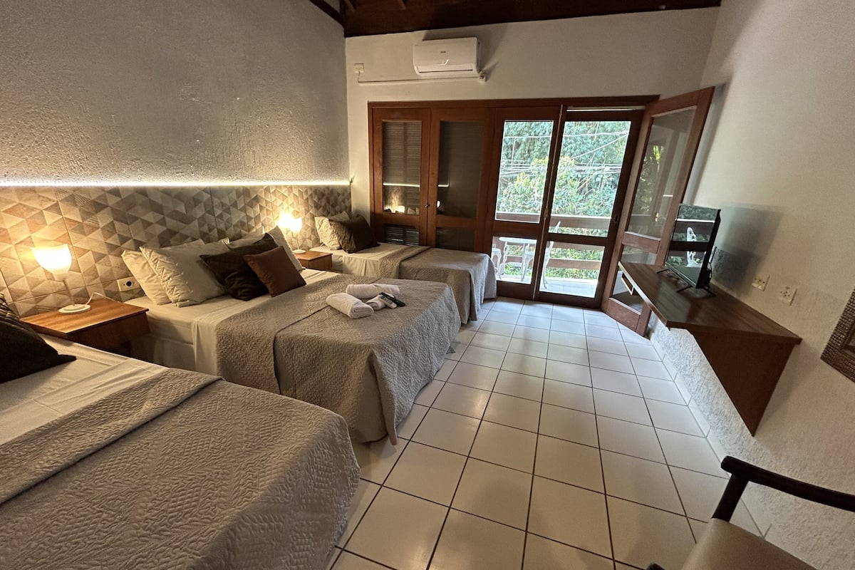 A hotel room with two beds, a TV, and a balcony with a view of greenery.