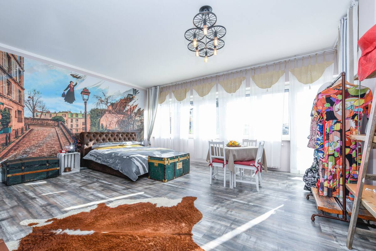 A bedroom with a large mural, a bed, a wooden floor, a chandelier, and a clothing rack.