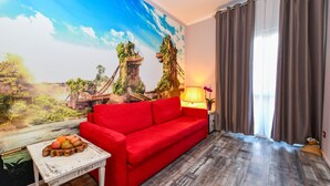 Ein Zimmer mit einer roten Couch, einem kleinen weißen Tisch mit Obst und einem Wandbild einer Brücke und viel Grün.