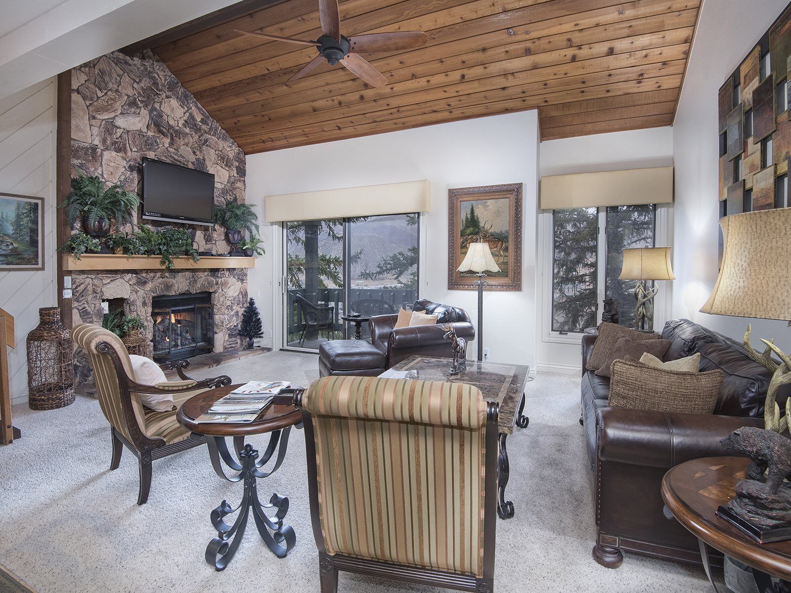 A spacious living room with a stone fireplace, leather furniture, and large windows offering a view of the outdoors.