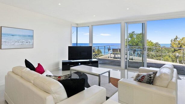 A modern living room with a flat-screen TV, white sofas, and a large window overlooking the sea.