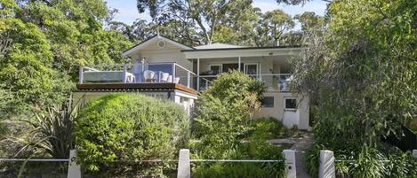 A two-story house with a balcony, surrounded by dense greenery and trees.