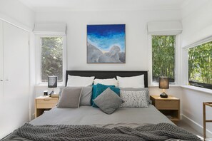 A bedroom with a bed, two bedside tables, a night lamp, a painting, and windows with a view of greenery.