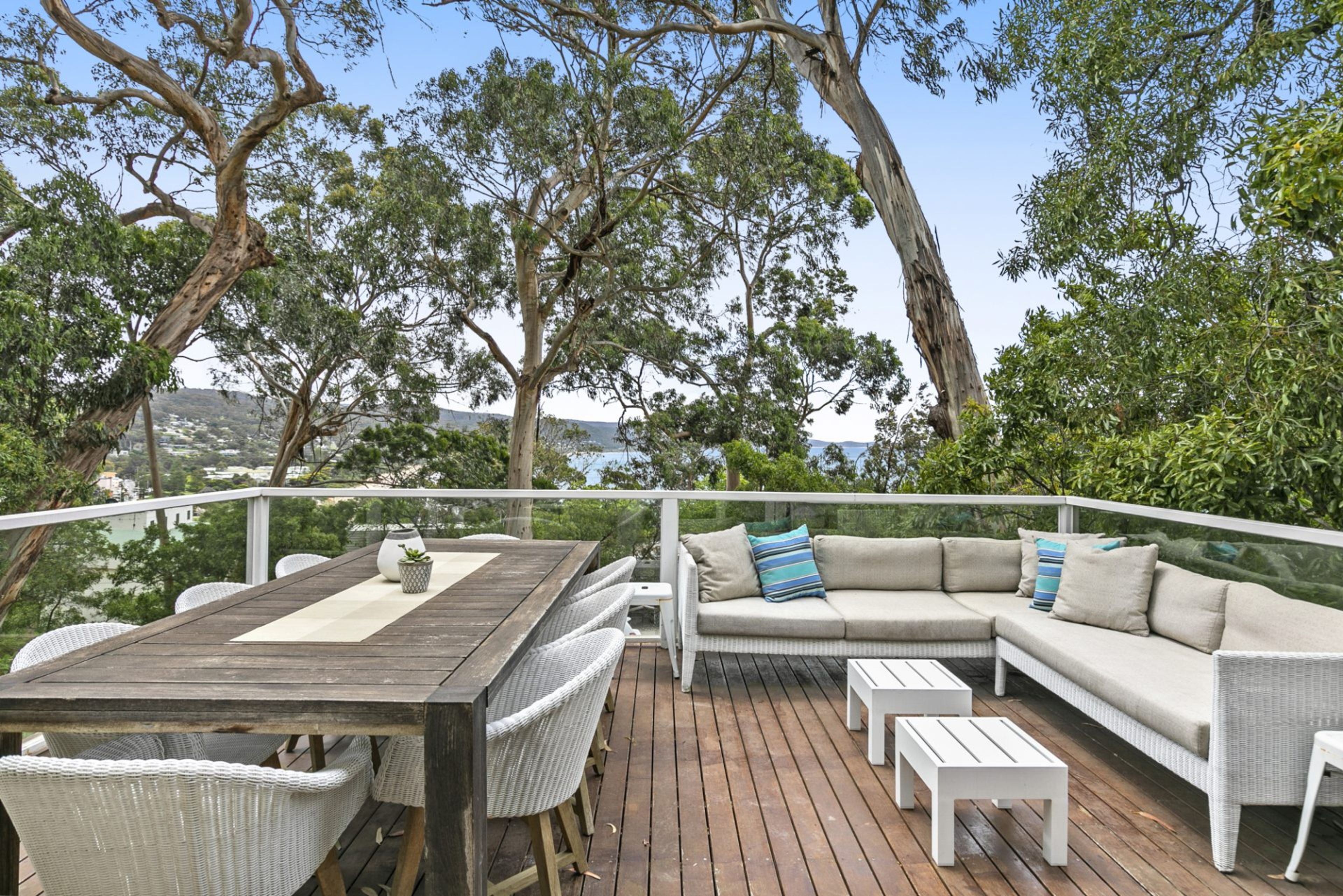 Eine Terrasse im Freien mit Holztisch, Korbstühle, einer Couch mit Kissen und einem Geländer mit Blick auf eine Landschaft mit Bäumen und weiter Aussicht.