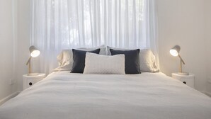 Una cama bien tendida con ropa de cama blanca, dos lámparas de noche y una ventana con cortinas traslúcidas.
