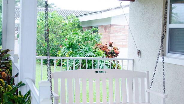 Eine Veranda mit einer weißen Schaukel, einem Deckenventilator und Blick auf einen Garten mit einer Ziegelmauer und einem Fenster.