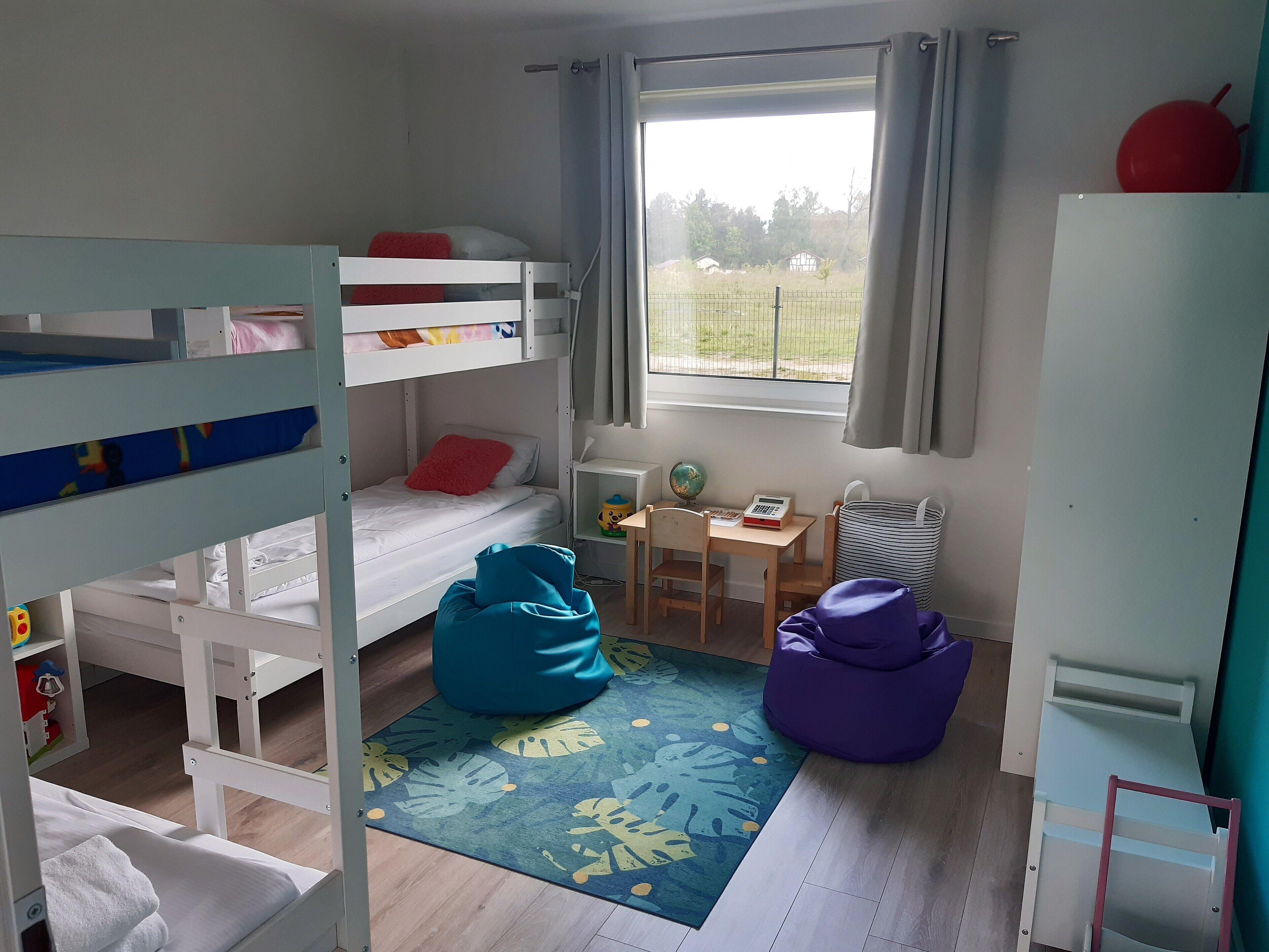 Ein Zimmer mit Etagenbetten, einem Schreibtisch, einem Stuhl, einem Globus, einem Rechner und einem Fenster mit Vorhängen.