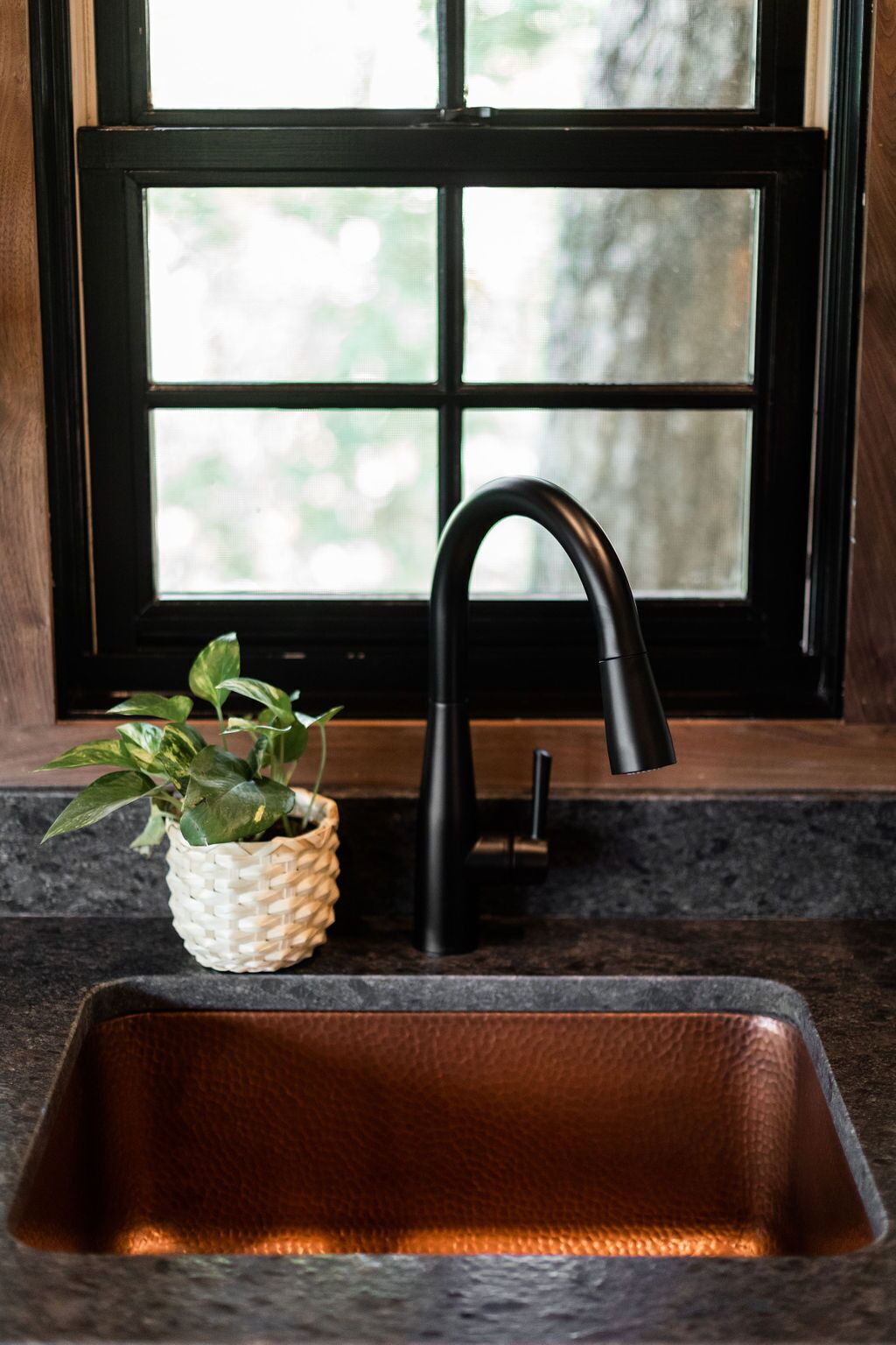 A kitchen sink with a black faucet, a woven plant pot, and a window with a view of greenery.