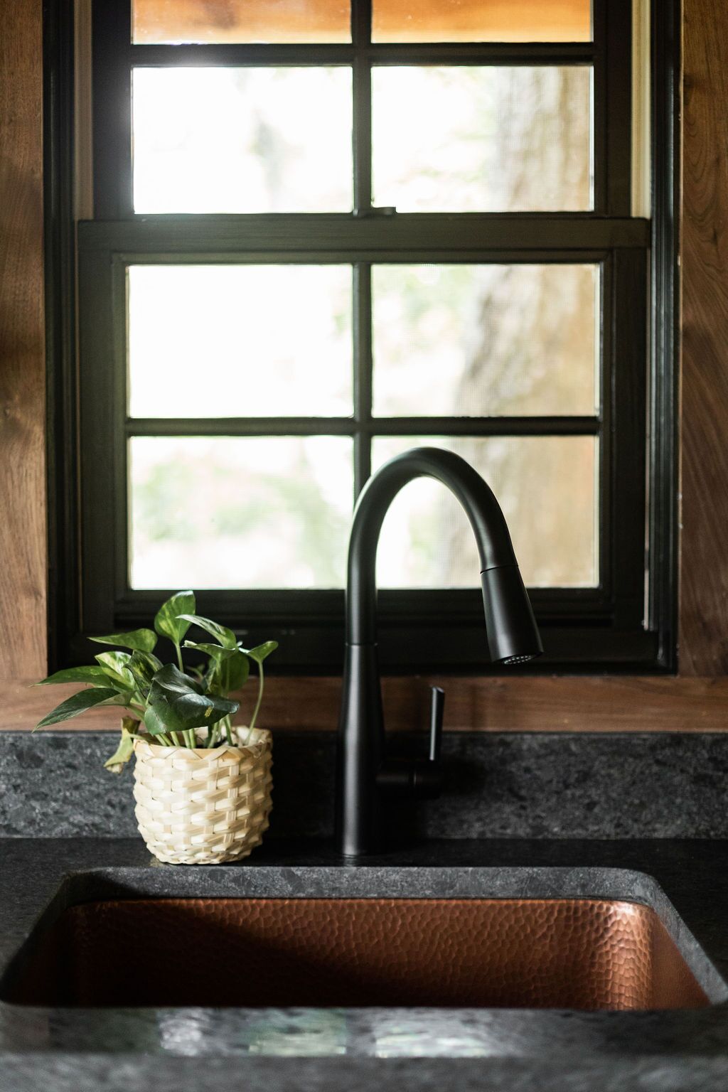 Un évier de cuisine avec un robinet noir, un panier tressé contenant des plantes vertes et un récipient d’évier en cuivre.