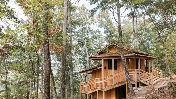 Een houten chalet met balkon, omringd door hoge bomen in een bos.