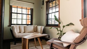 Een gezellige kamer met een bank, een stoel, een houten tafel en grote ramen met luxaflex.