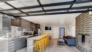 A modern kitchen with a central island, yellow chairs, a grey sofa, and a brick fireplace.