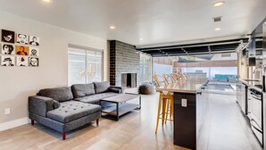 A modern kitchen with a central island, a dining area with chairs, and a grey sectional sofa.