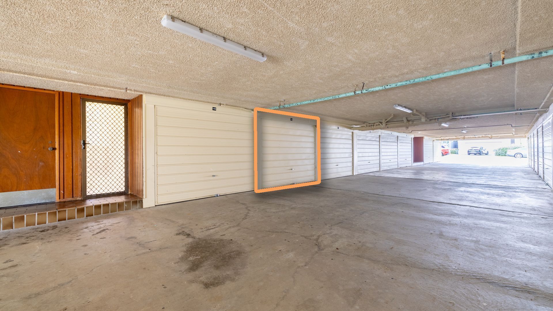 Een lange, lege parkeergarage met betonnen vloeren en muren, meerdere gesloten garagepoorten en een houten deur met hor.
