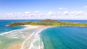 A coastal landscape with a sandy beach, clear blue waters, and a hilly island with dense vegetation.