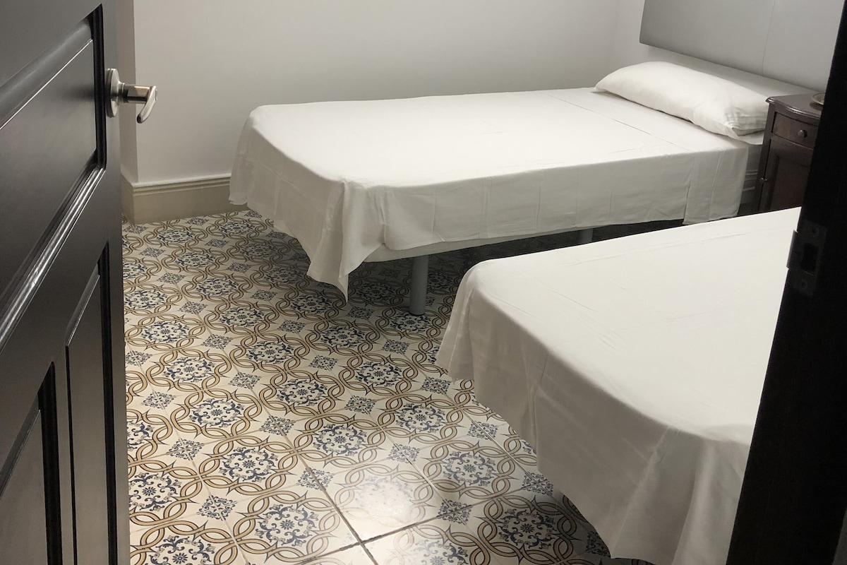 A small, clean room with two single beds, a round ceiling light, and patterned floor tiles.
